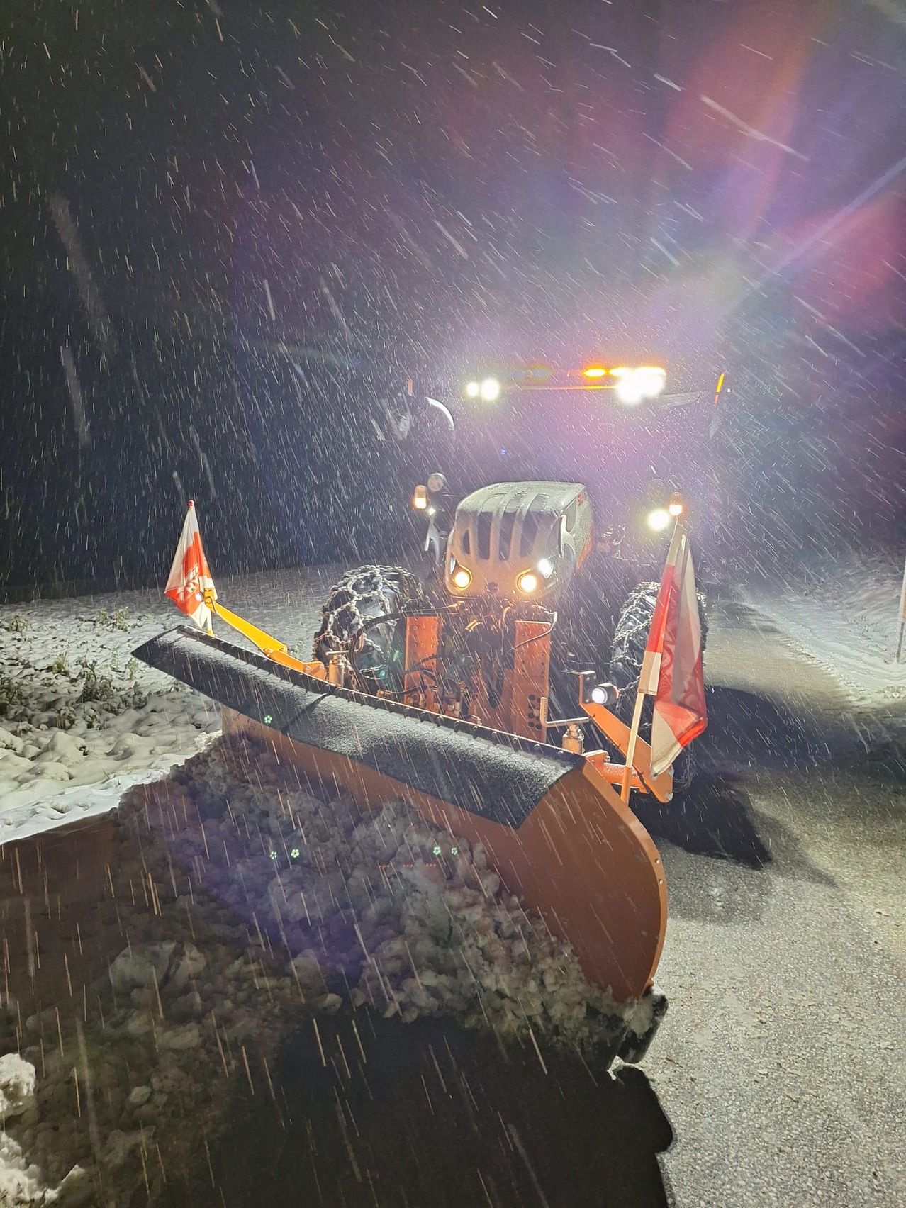 Ein Schneepflug räumt bei Nacht eine verschneite Straße. Er hat zwei Flaggen vorne und seine Scheinwerfer sind eingeschaltet.