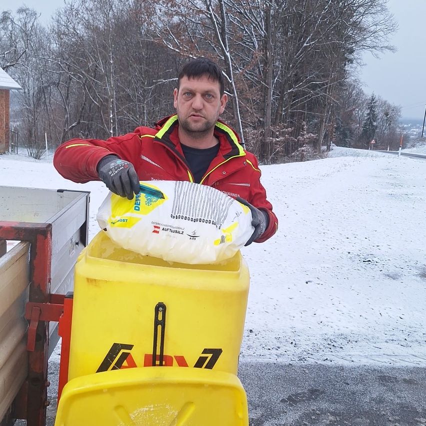 Ein Mann in einem roten Mantel und Handschuhen hält ein weißes Paket vor einem gelben Mülleimer auf einer verschneiten Straße.