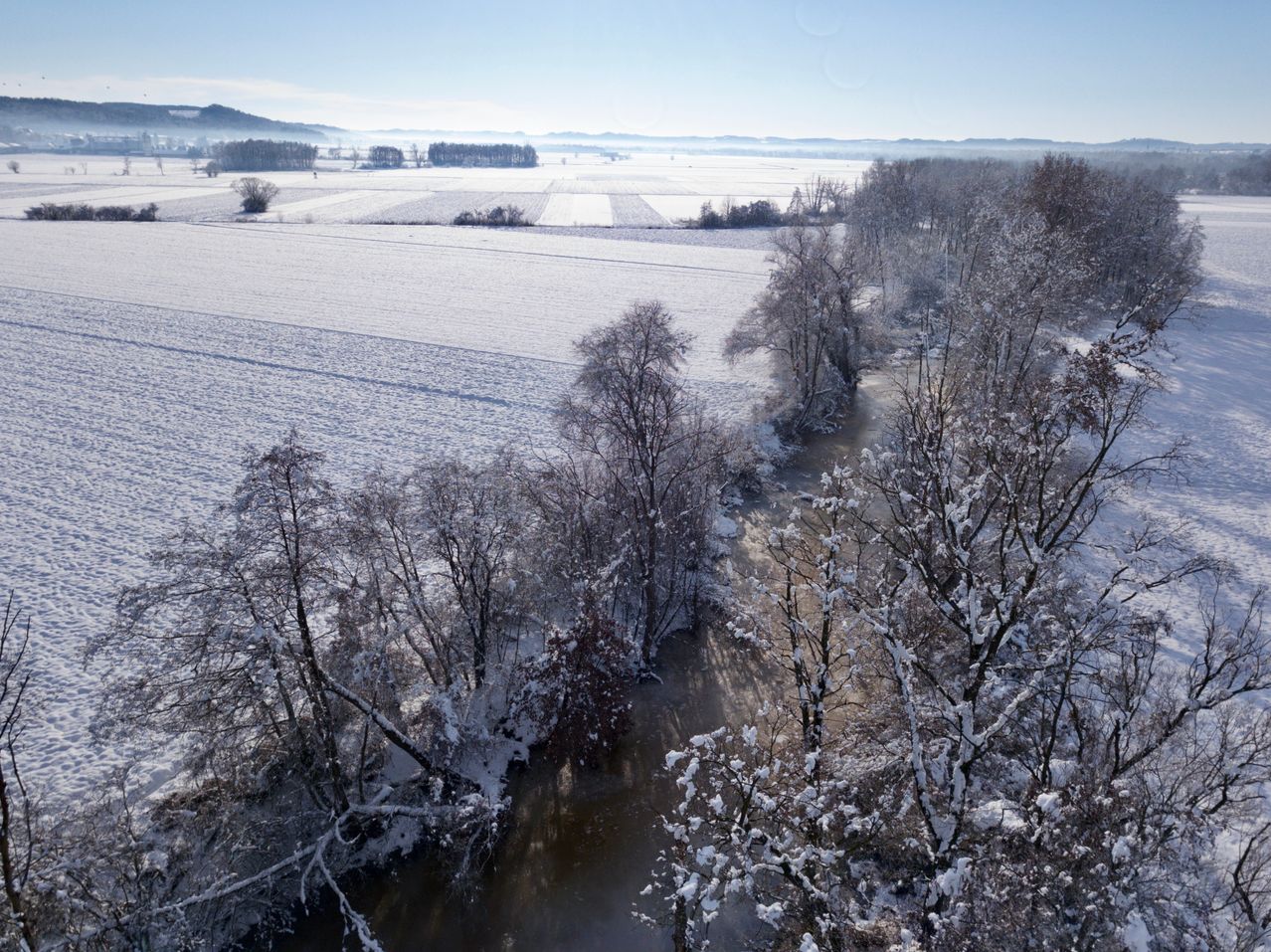 Luftaufnahme einer verschneiten Landschaft mit Bäumen, einem Fluss und kahlen Feldern. Der Himmel ist klar.