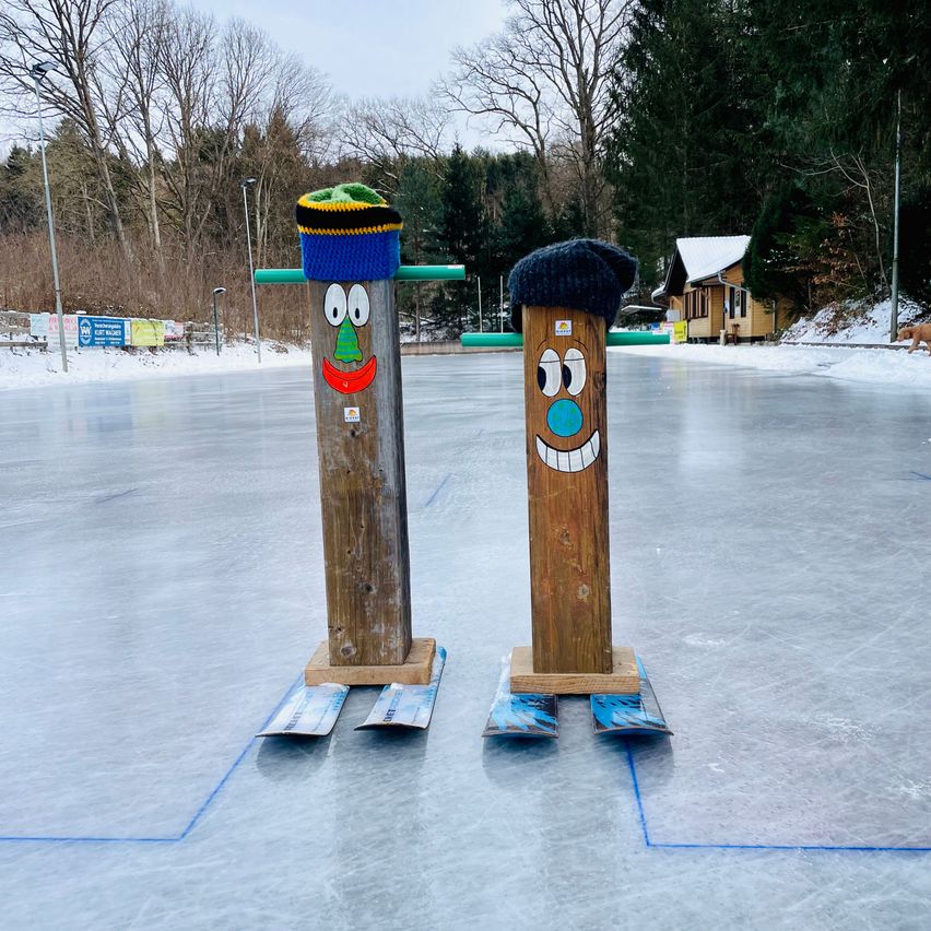 Zwei Holzpfosten mit bemalten Gesichtern stehen auf einer verschneiten Eisbahn, mit einem Haus und Bäumen im Hintergrund.