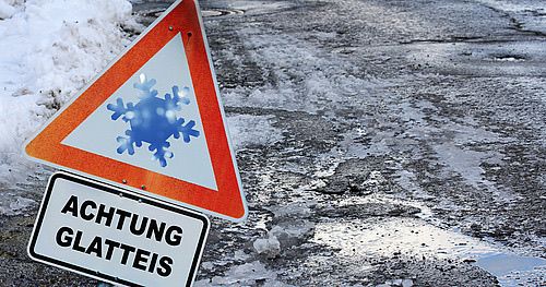 Ein dreieckiges Schild mit einem Schneeflockensymbol warnt vor schwarzem Eis auf der Straße. Darunter befindet sich ein rechteckiges Schild mit der Aufschrift 'ACHTUNG GLATTIEIS' auf Deutsch.