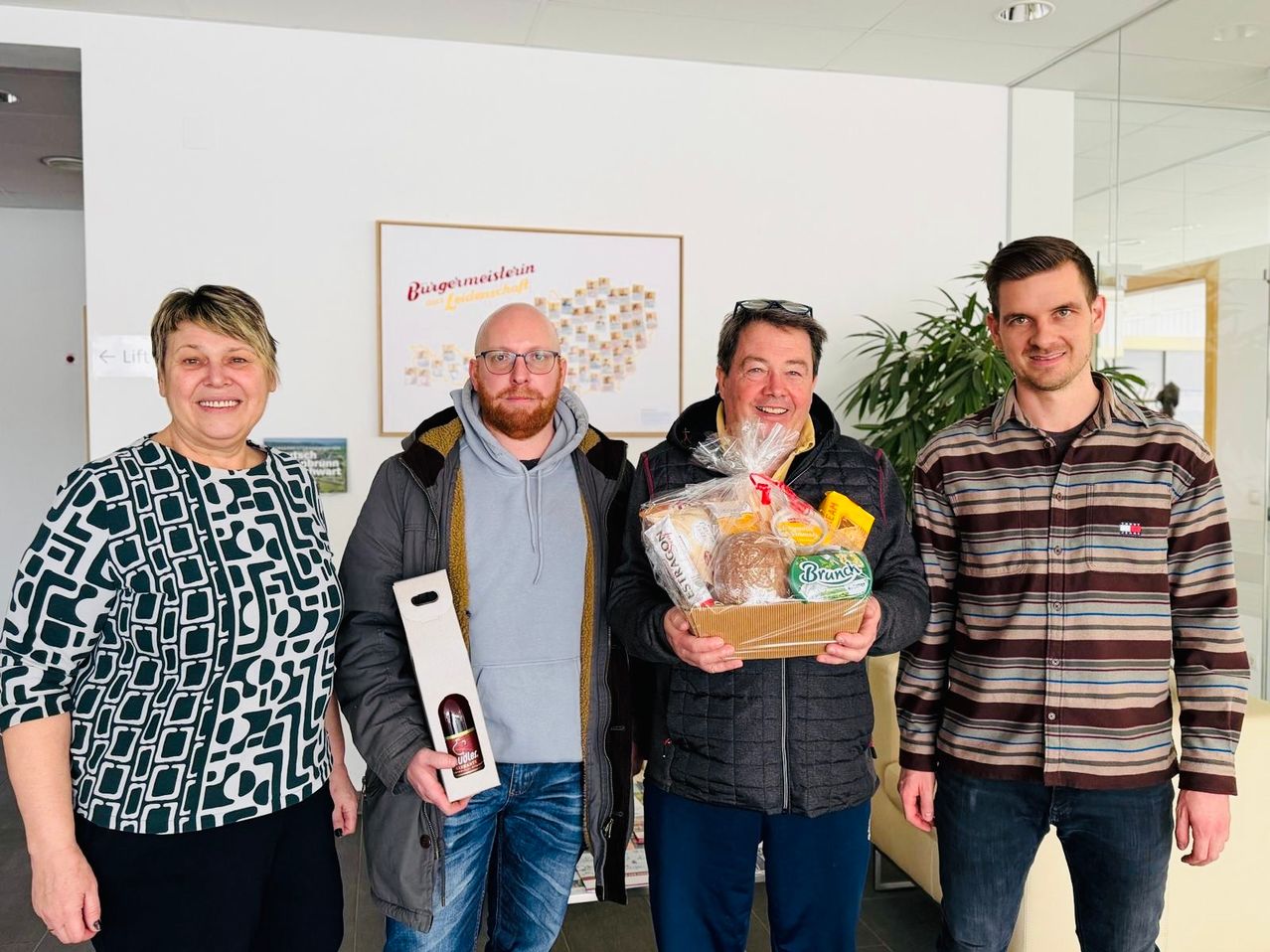 Vier Menschen stehen eng beieinander im Innenraum. Ein Mann hält eine Box mit Essen und ein anderer eine Flasche. Der Mann in der Mitte trägt eine Brille und eine Sonnenbrille. Hinter ihnen ist ein Bild und eine Pflanze.