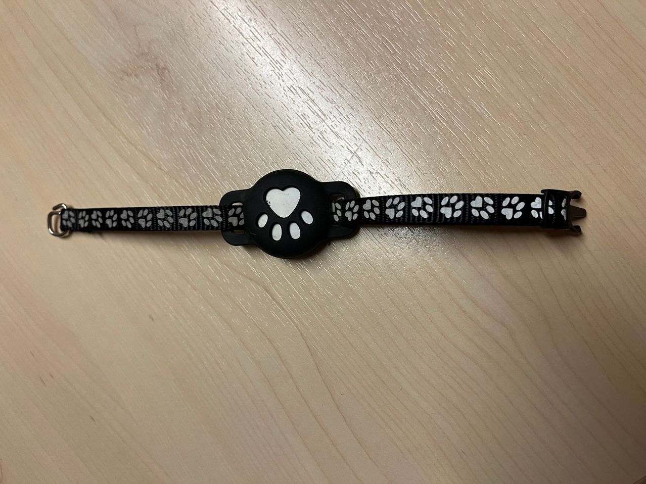A black and white paw print collar with a heart buckle lies flat on a wooden surface.