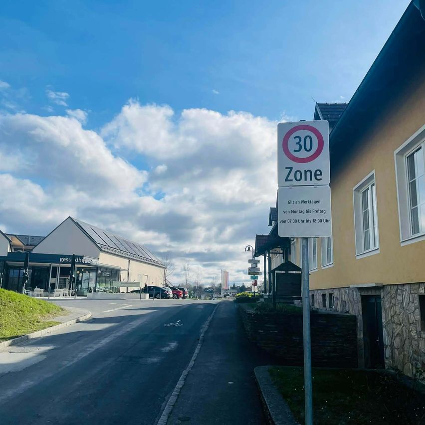 Ein 30er-Zonenschild an einem Pfosten neben der Straße. Ein Gebäude mit Solarpanelen im Hintergrund. Der Himmel ist blau mit Wolken.
