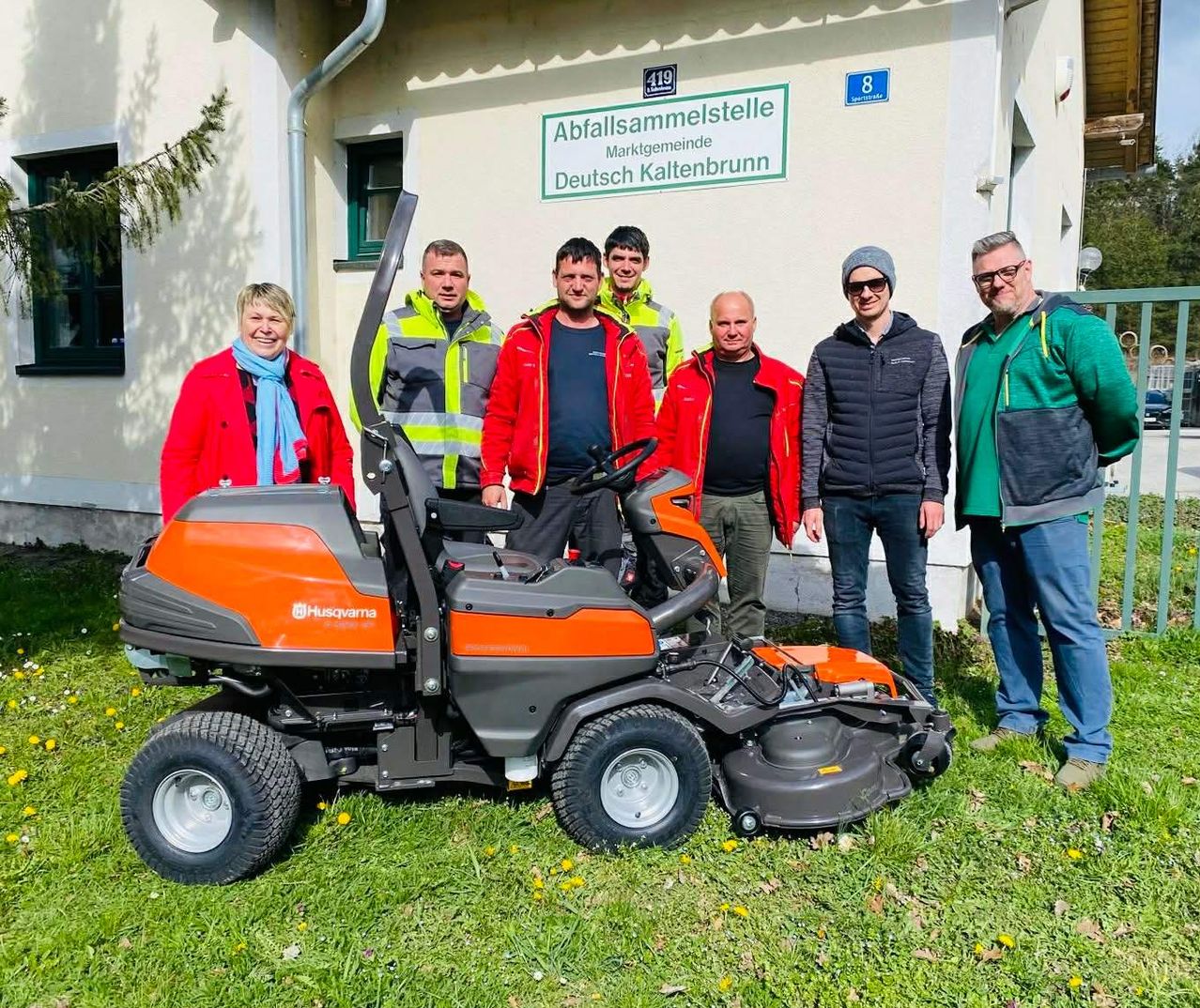 Eine Gruppe von Menschen steht vor einem Gebäude mit der Aufschrift Abfallsammlungsstelle Marktgemeinde Deutsch Kaltenbrunn. Sie posieren mit einem Husqvarna-Rasenmäher.