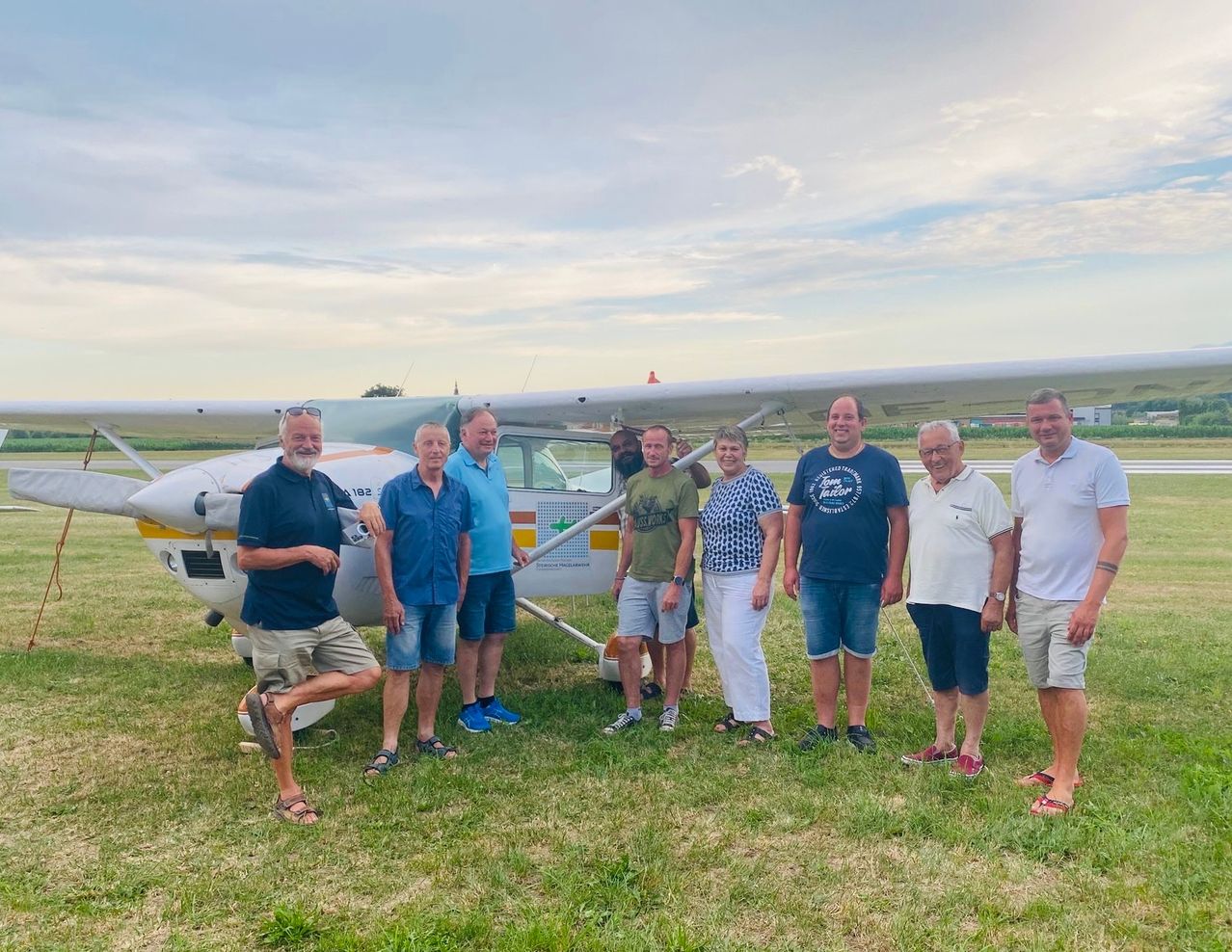 Acht Personen posieren für ein Foto vor einem weißen Flugzeug auf einem Rasenfeld.