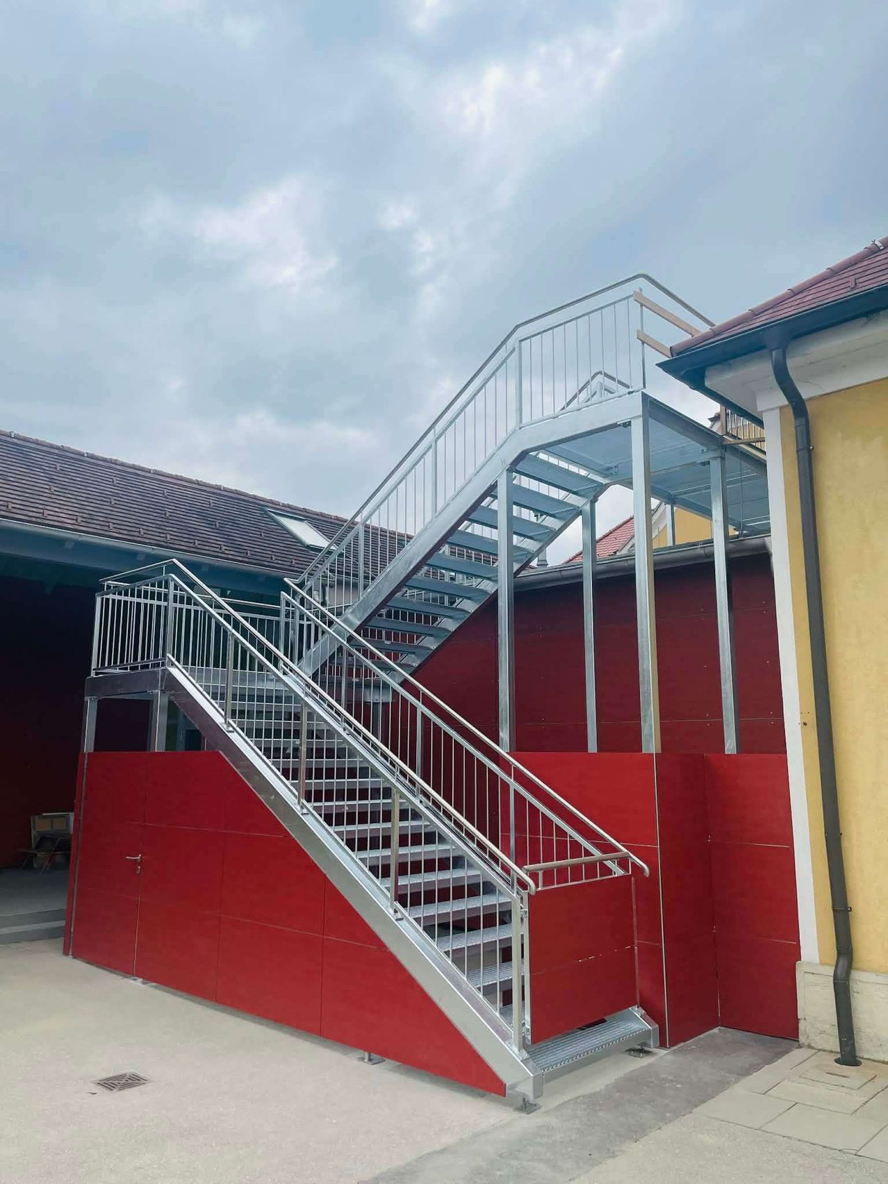 Eine Außentreppe aus Metall mit Handläufen an einer roten Wand und einem grauen Dach an einem bewölkten Tag.