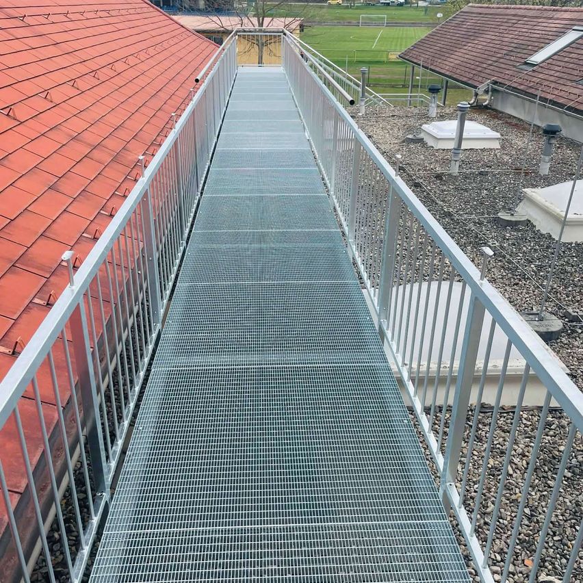 Ein Stahlgitterweg mit Metallgeländern auf beiden Seiten, der zwei Gebäude verbindet. Der Weg ist im Freien mit Kies an den Seiten.