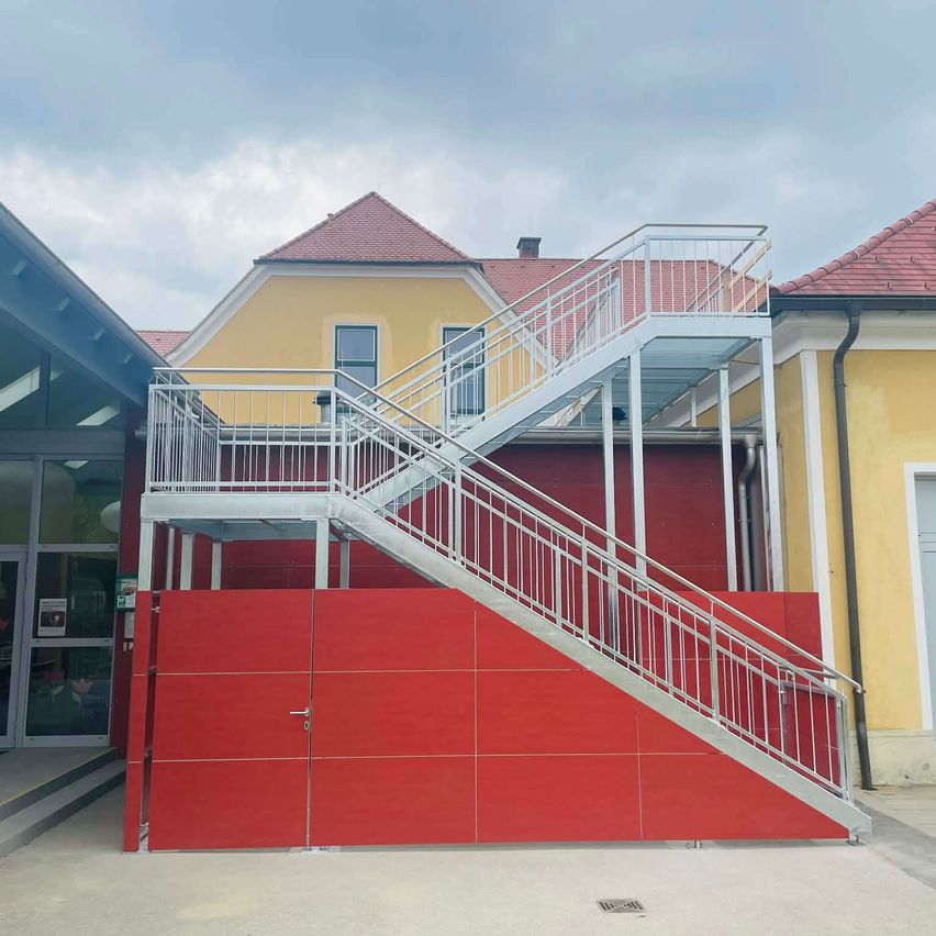 Ein Gebäude mit einer roten Wand, einer weißen Treppe und einem roten Dach. Die Wand hat zwei Schiebetüren, und die Treppe hat ein Metallgeländer. Der Himmel ist bewölkt.