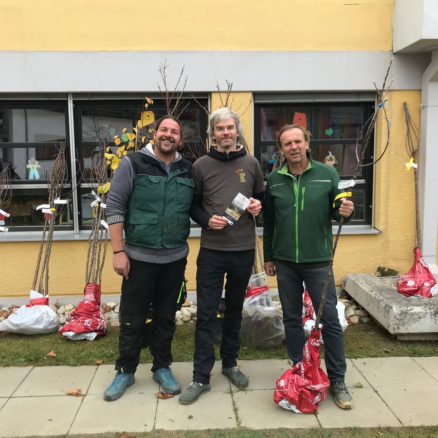 Three men stand outside near a building, smiling. One holds a pamphlet, and they all hold sticks. They have bags of plants behind them.