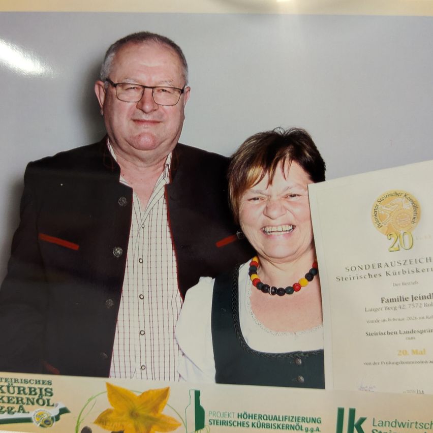 An older couple stands together smiling. The man wears glasses and a suit with a white shirt. The woman wears a necklace and a traditional dress. They hold a certificate with the words 'Sonderauszeihung Steirisches Kürbiskernöl'.