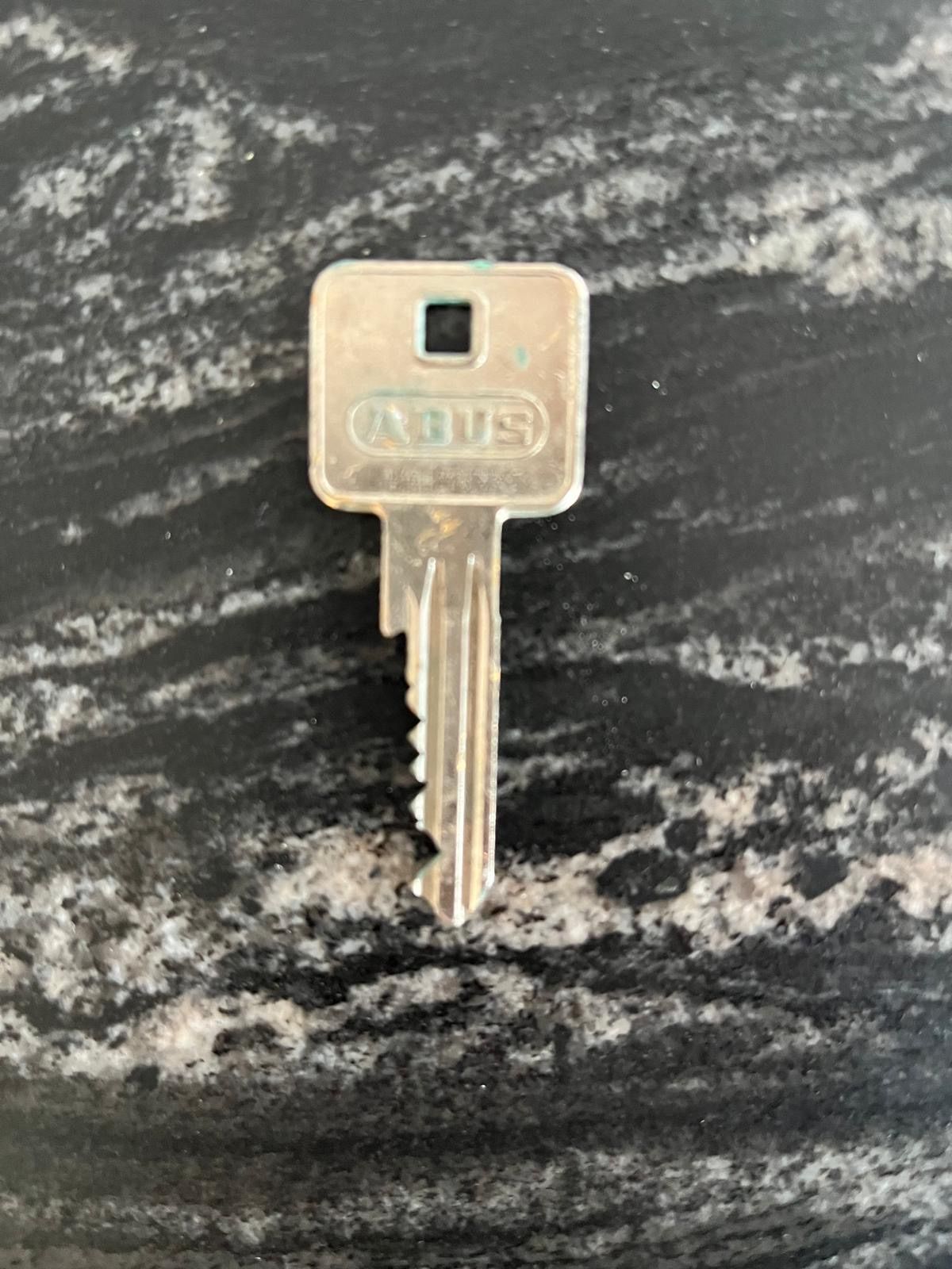 A silver key with a square head and teeth, labeled ABUS, rests on a black marble surface.
