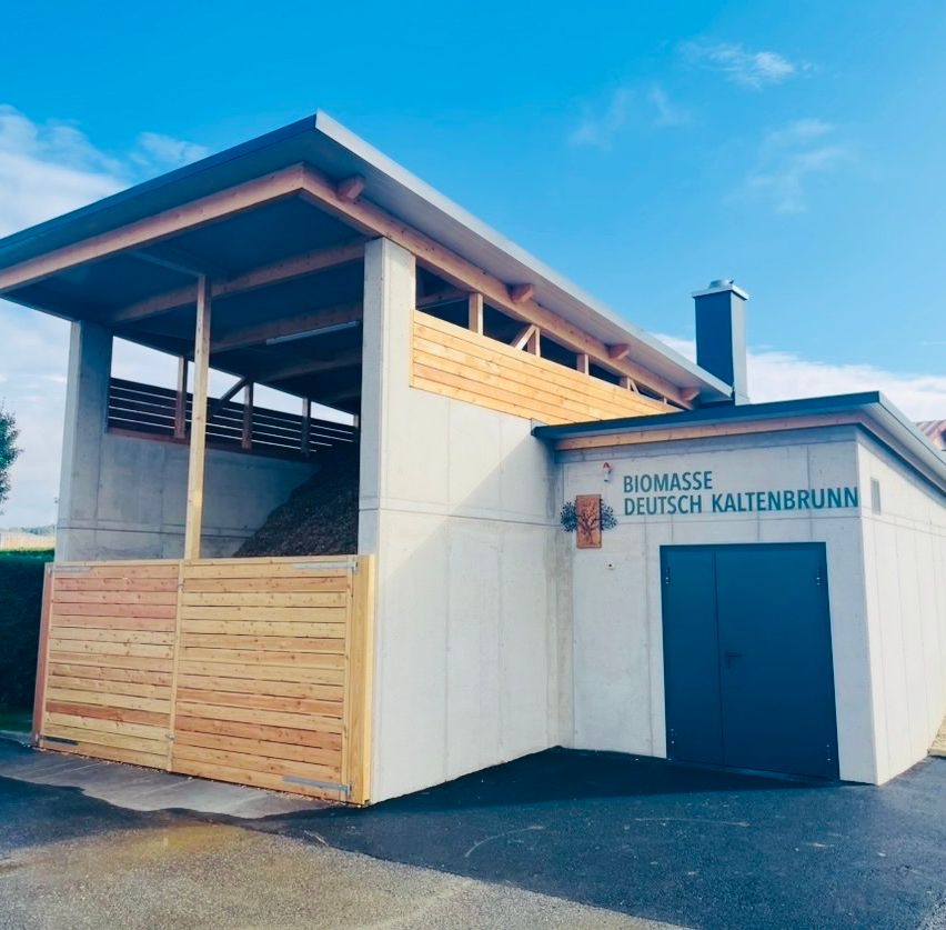 Ein kleines, modernes Gebäude mit einem Holzdach und einer großen Materialhaufen im Inneren. Das Gebäude trägt ein Schild mit der Aufschrift 'Biomasse Deutsch Kaltenbrunn'.