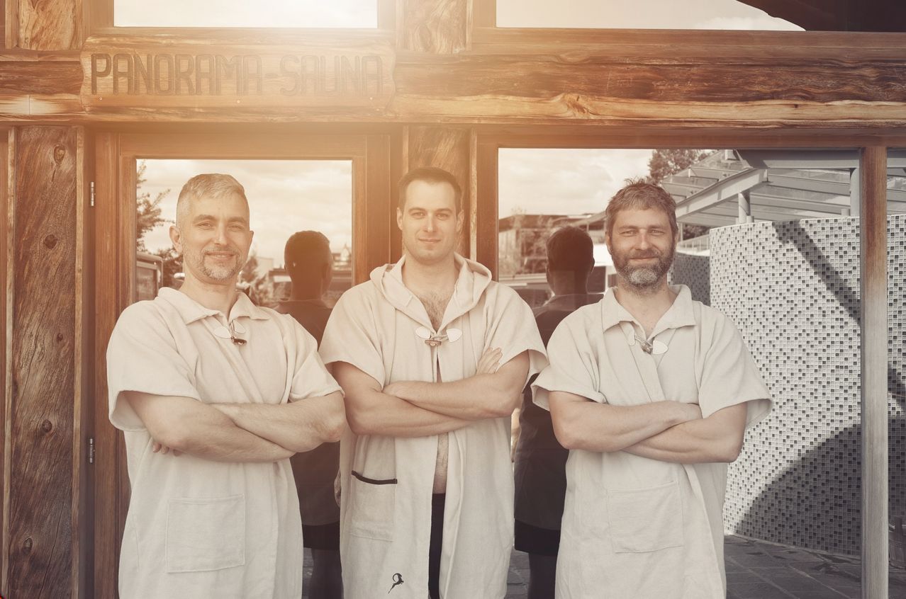 Drei Männer in weißen Roben stehen vor einem Holzschild, das 'Drama Sauna' besagt. Sie lächeln und haben die Arme verschränkt. Hinter ihnen sind zwei Personen durch die Glastür teilweise sichtbar.