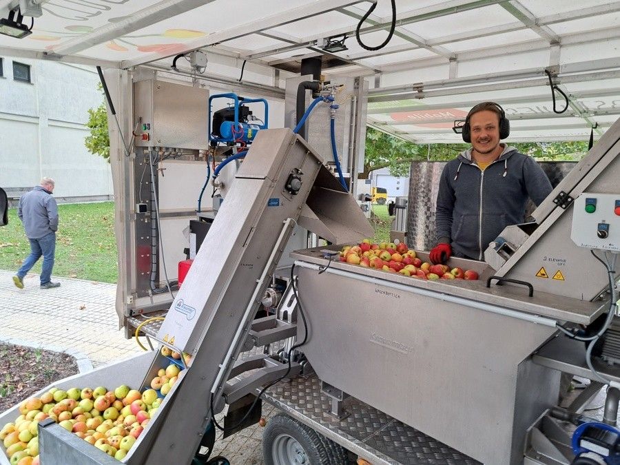Ein Mann mit Kopfhörern und einer grauen Kapuzenjacke bedient eine Maschine zur Verarbeitung von Äpfeln, wobei ein Förderband und Edelstahlbehälter sichtbar sind.