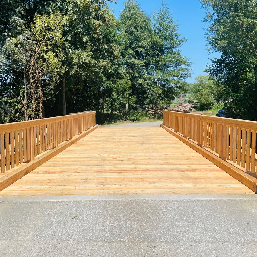 Eine hölzerne Brücke mit Geländern erstreckt sich über eine Straße, umgeben von üppigem grünem Laub und einem klaren blauen Himmel.
