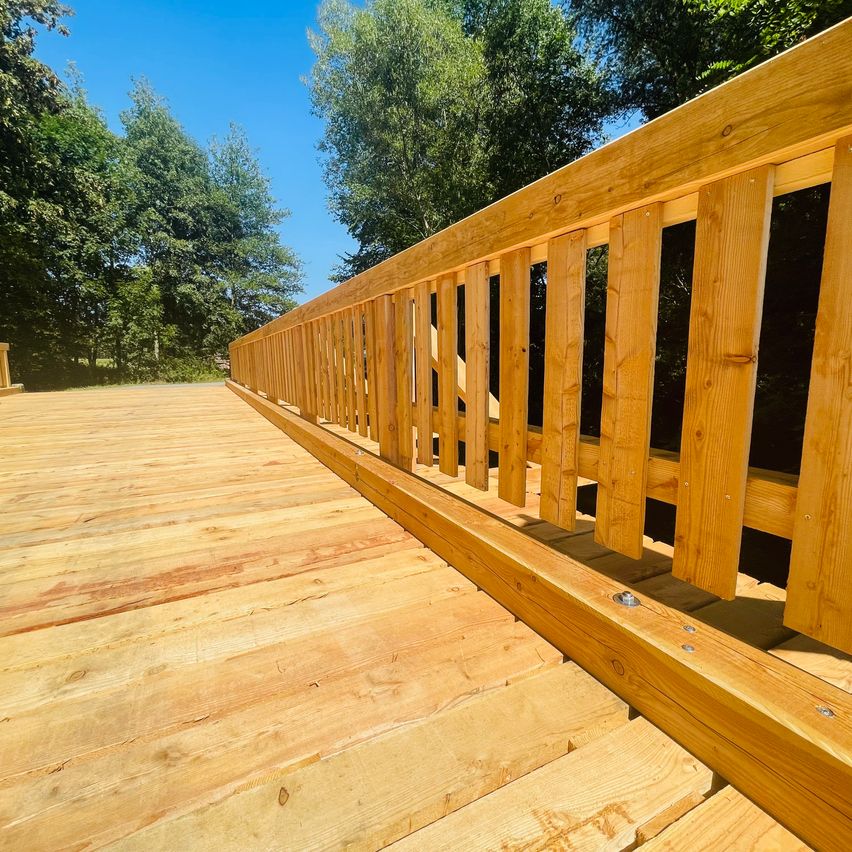 Holzbrücke mit Geländer und Lattenzaun vor Bäumen und blauem Himmel