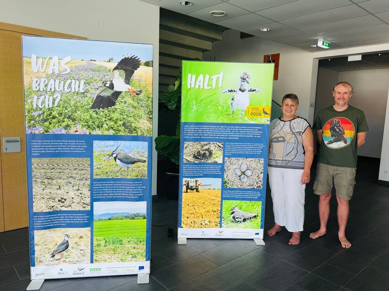 Drei Personen stehen neben Postern mit Vögeln und Text. Die Frau und der Mann rechts lächeln in die Kamera. Das Poster links zeigt einen Vogel, der über ein Feld fliegt. Das mittlere Poster zeigt einen Vogel auf Gras mit dem Text 'HALTI'. Das rechte Poster zeigt einen Vogel in einem Nest mit Eiern.