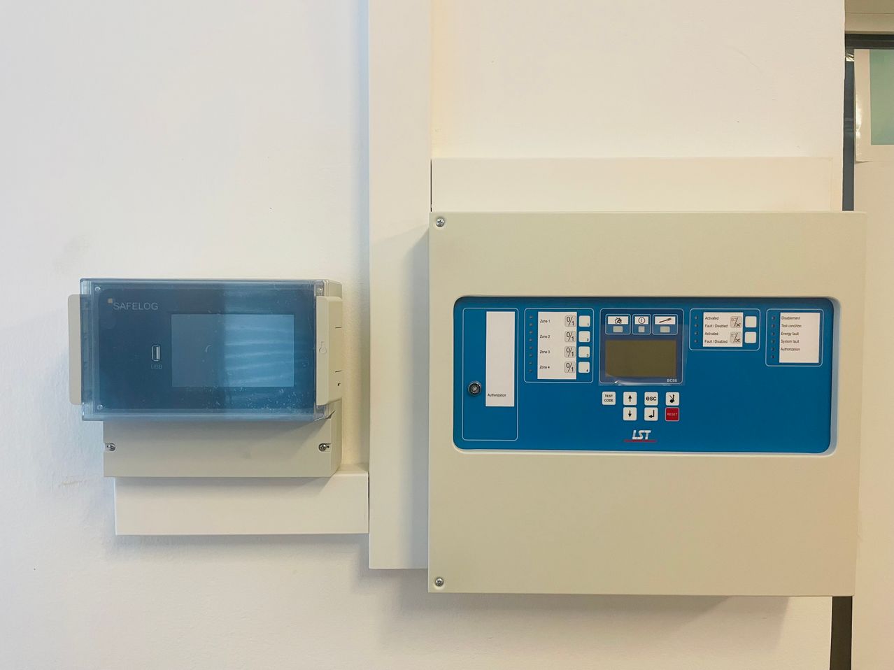 Zwei elektronische Geräte an der Wand montiert. Das linke Gerät ist mit Safelog beschriftet, und das rechte Gerät hat eine blau-weiße Schnittstelle mit Tasten und einem Bildschirm.