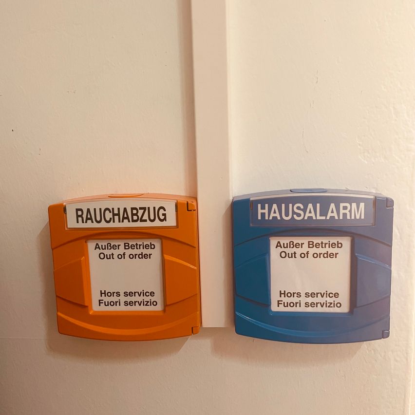 Zwei Rauchmelderabdeckungen sind an einer weißen Wand montiert. Die linke Abdeckung ist orange mit der Aufschrift 'RAUCHABZUG' und 'Außer Betrieb'. Die rechte Abdeckung ist blau mit der Aufschrift 'HAUSALARM' und 'Außer Betrieb'. Beide haben einen Serviceaufkleber 'Hors service Fuori servizio'.