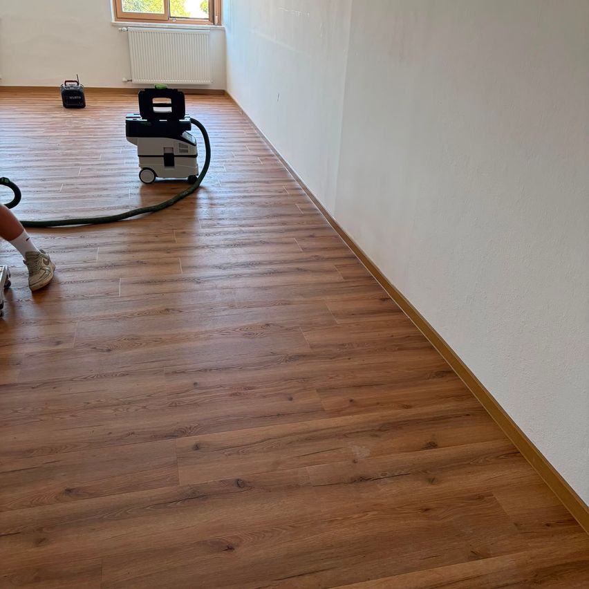 Eine Person saugt einen Holzboden in einem leeren Raum mit weißen Wänden und einem Fenster.