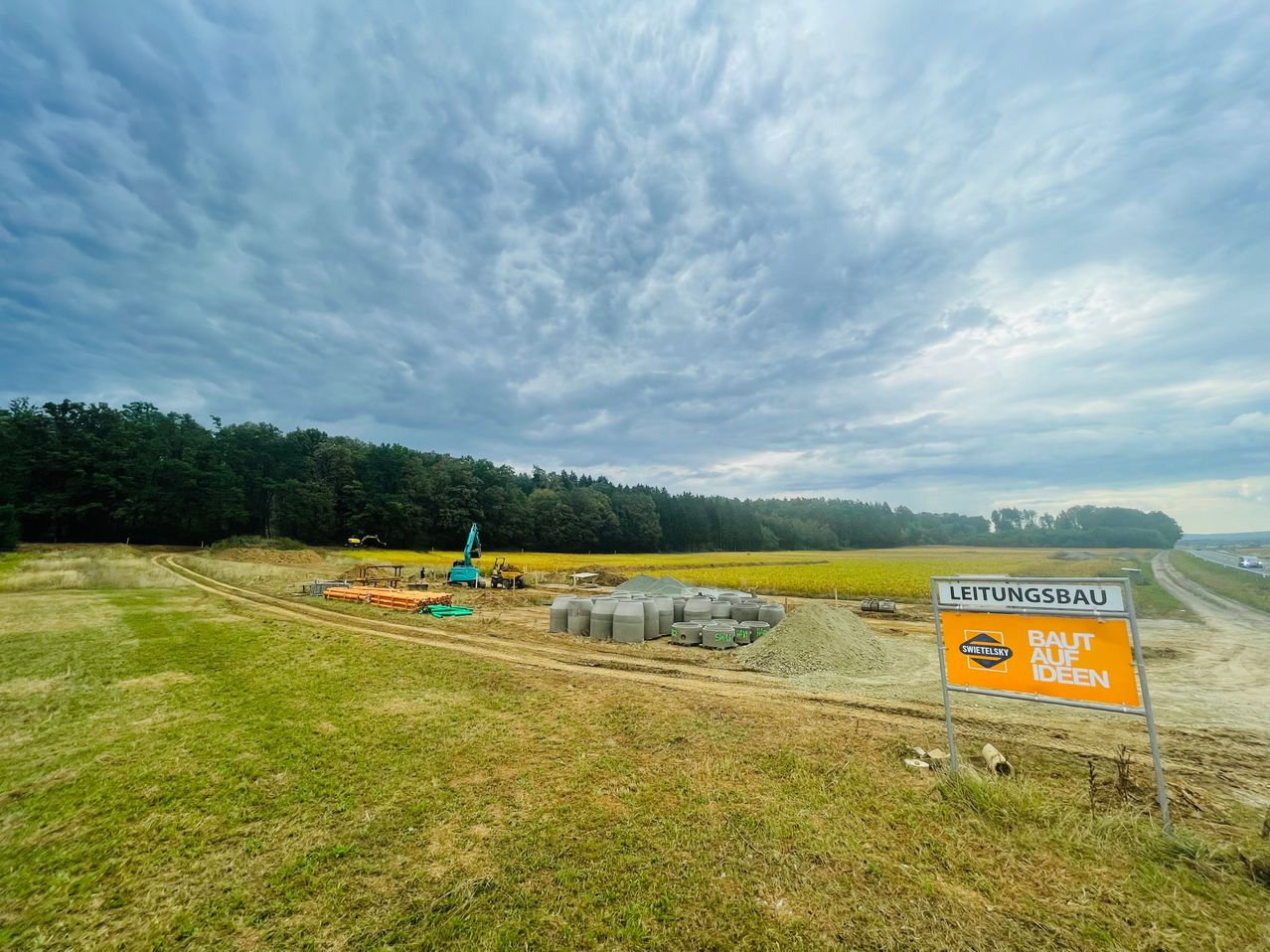 Eine Baustelle mit einem Schild, das 'Leitungsbau' und 'Baut auf Ideen' besagt. Maschinen und Materialien sind verstreut. Bäume und ein bewölkter Himmel im Hintergrund.