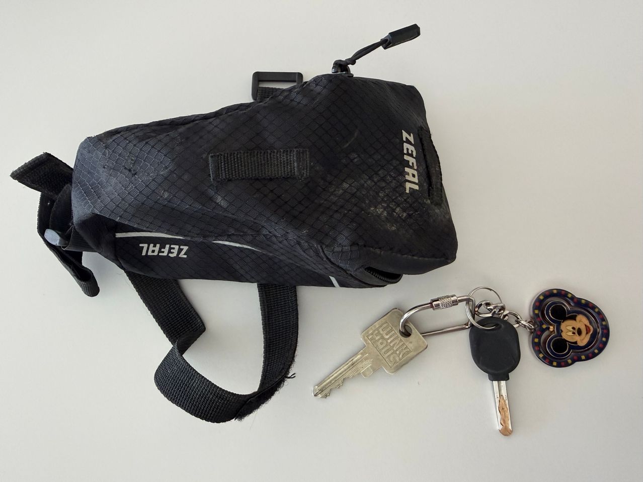A black Zefal bag with a strap and zipper, alongside two keys, one silver and one black, on a white surface.