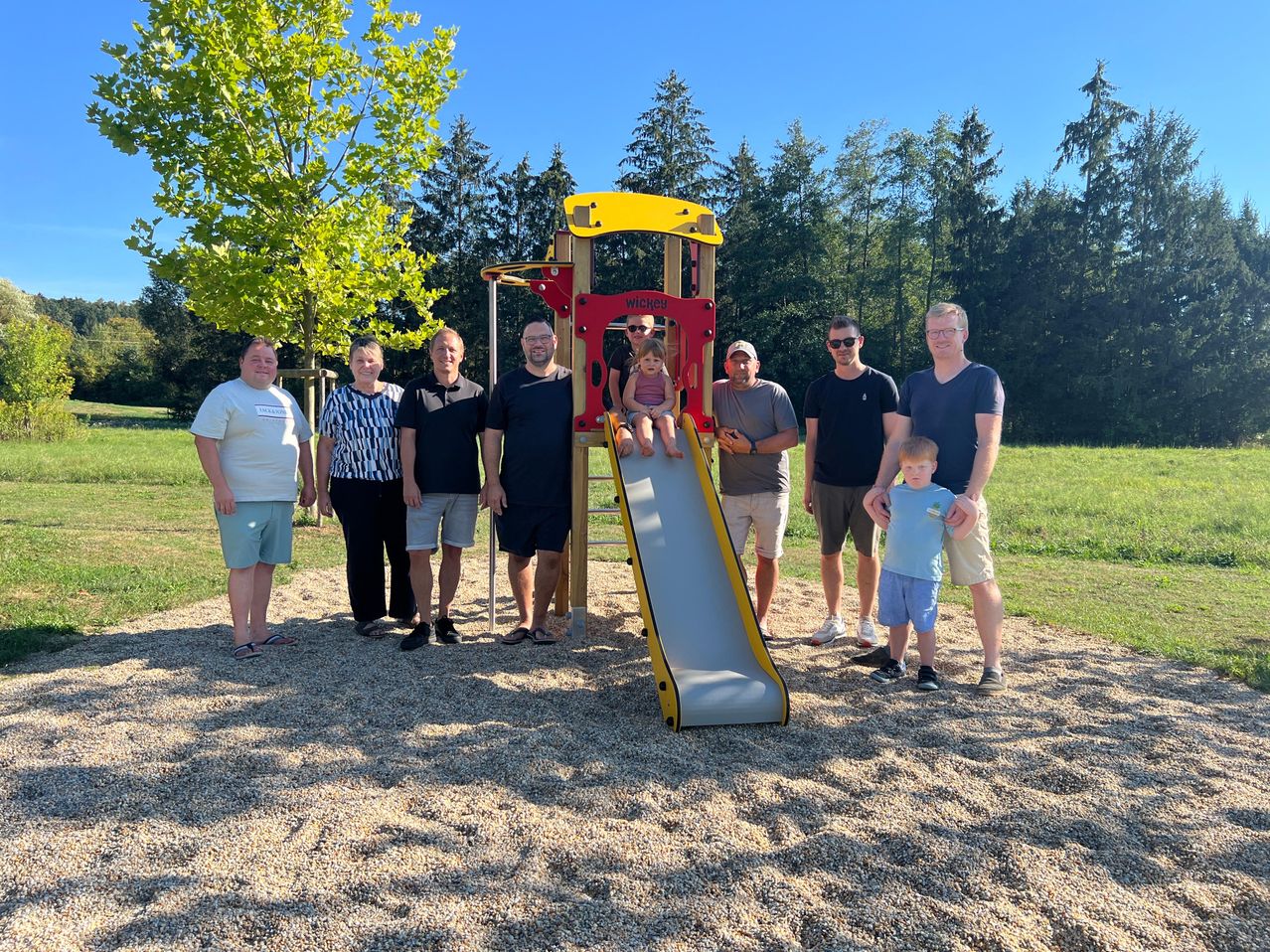 A group of people, including adults and children, are posing for a photo at a playground. A young girl is climbing up the slide.