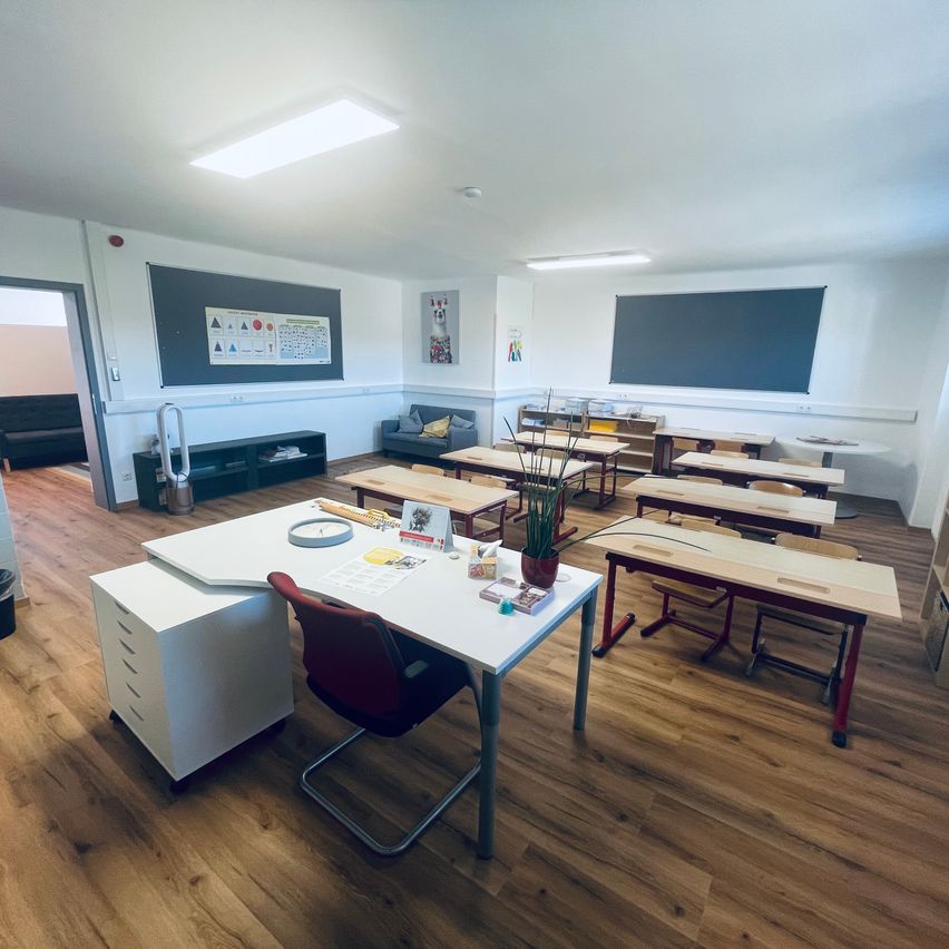 An empty classroom with wooden floors, white walls, and a teacher's desk at the front. There are rows of desks and chairs, and two blackboards on the wall.