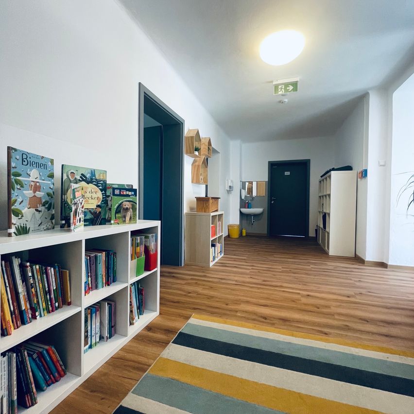 A well-lit room with wooden floors, white walls, and a bookshelf filled with children's books. There is a rug on the floor, a sink in the hallway, and a door leading to another room.