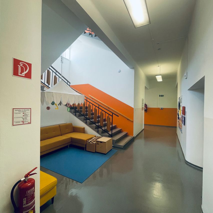 An empty hallway with a blue rug, a couch, and a fire extinguisher. The hallway has white walls, an orange staircase, and a white tent.