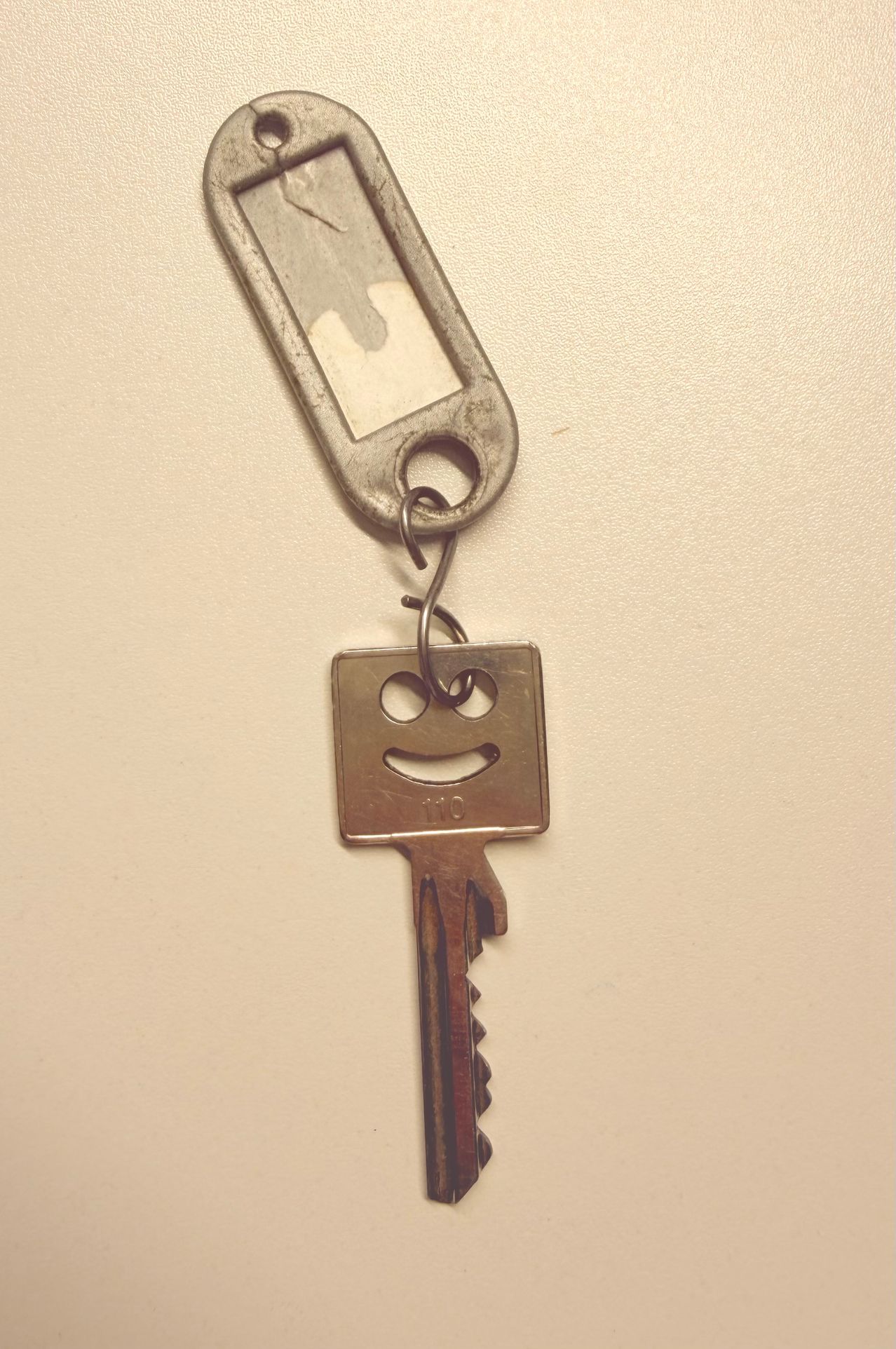 Ein Schlüssel mit einem eingeätzten lächelnden Gesicht, befestigt an einem Schlüsselbund mit einem kleinen, abgenutzten Etikett.