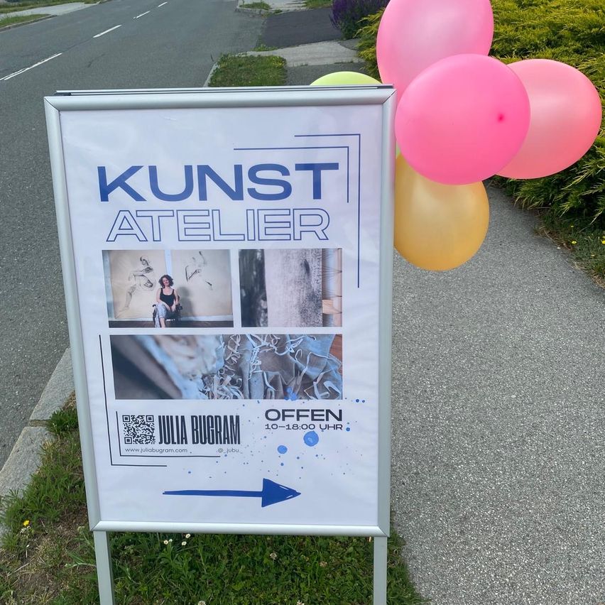 Ein Schild für Kunst Atelier, das verschiedene Kunstwerke und Öffnungszeiten zeigt. Rosa und gelbe Luftballons sind oben gebunden. Ein Pfeil zeigt nach rechts.