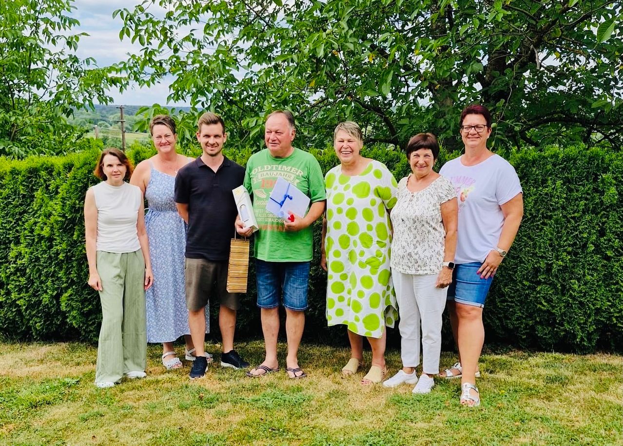 Sieben Personen stehen im Freien vor einer Hecke und Bäumen.