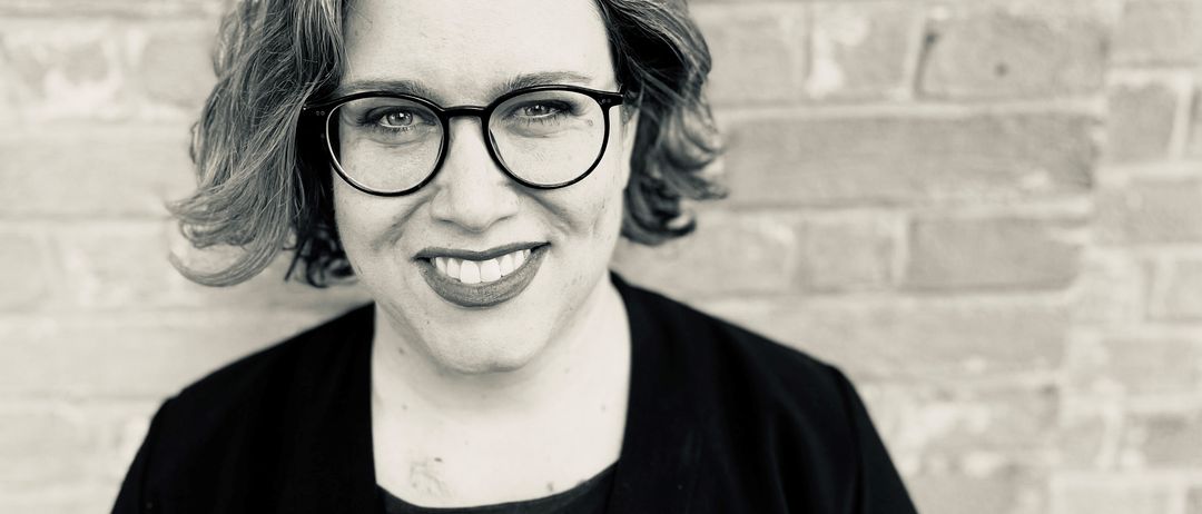 Schwarz-Weiß-Porträt einer lächelnden Frau mit Brille und schwarzem Blazer vor einer Ziegelwand.