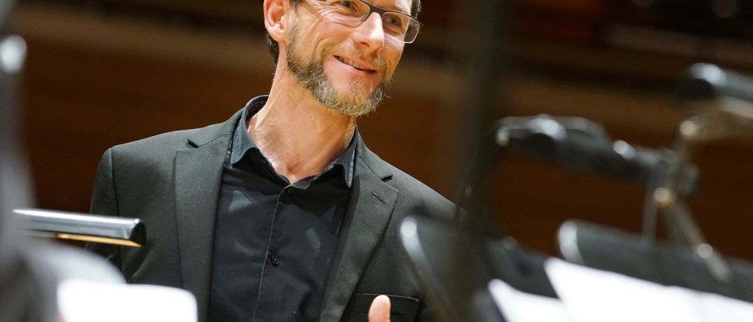 Ein lächelnder Mann in einem schwarzen Anzug, der einen Daumen hochgibt. Er trägt eine Brille und steht in einem Raum.