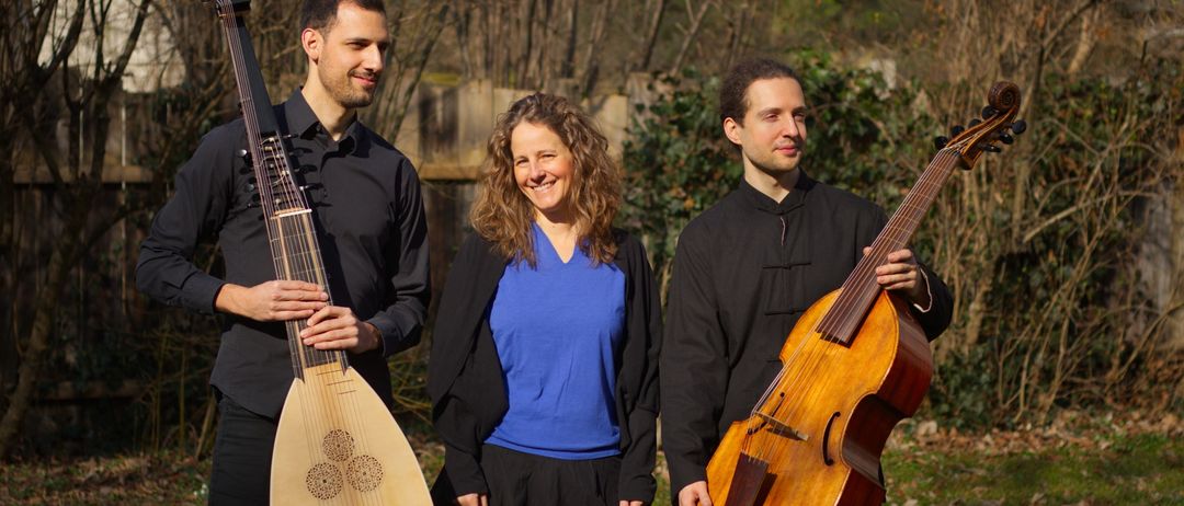 Drei Menschen stehen lächelnd zusammen in einem Außenbereich. Eine Frau in der Mitte trägt ein blaues Shirt, und zwei Männer an den Seiten halten eine Laute und ein Cello.