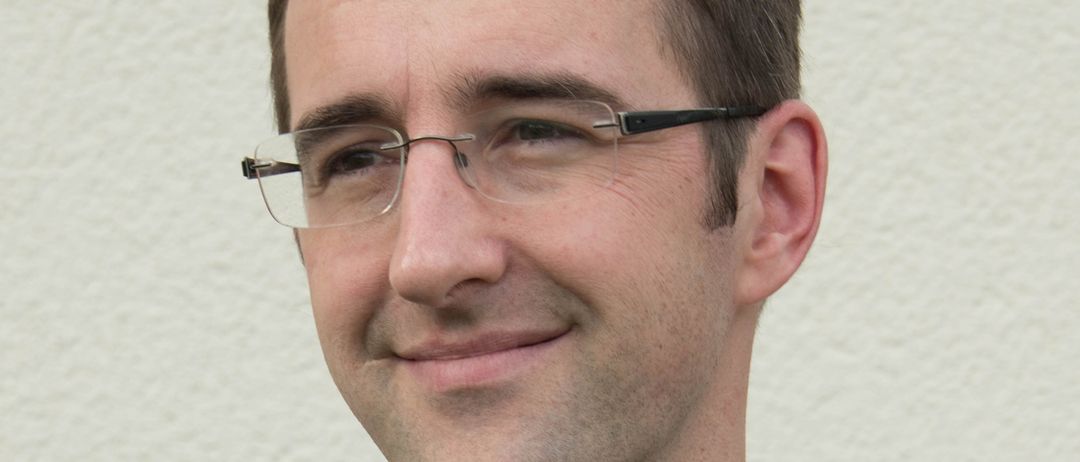 Ein Mann mit Brille in einem schwarzen Hemd, er lächelt und schaut in die Kamera.