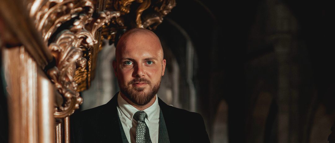 Ein Mann in formeller Kleidung steht in einem verzierten Flur, möglicherweise in einem historischen Gebäude. Er hat einen Bart und sieht zur Seite.