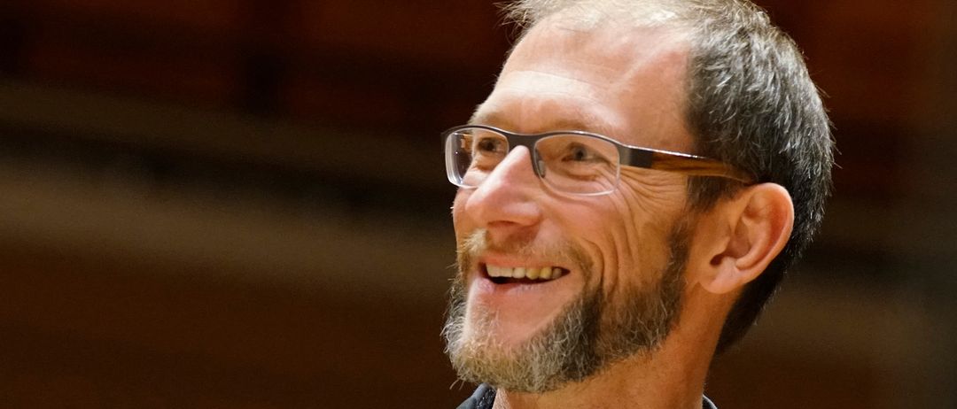 Ein Mann mit Brille und Bart lächelt und blickt nach rechts, möglicherweise in einer Aufführungssituation.