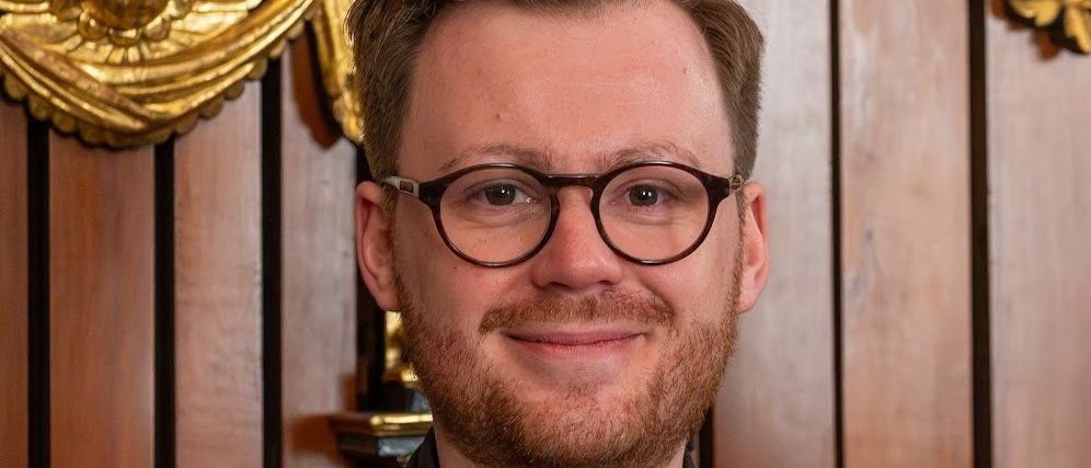 Ein Mann mit Brille, Bart und schwarzem Anzug steht lächelnd vor einer hölzernen Wand mit goldenen Verzierungen.