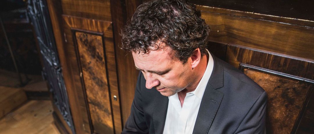 Ein Mann in einem Anzug spielt ein Musikinstrument, während er auf einer Holzbank sitzt.