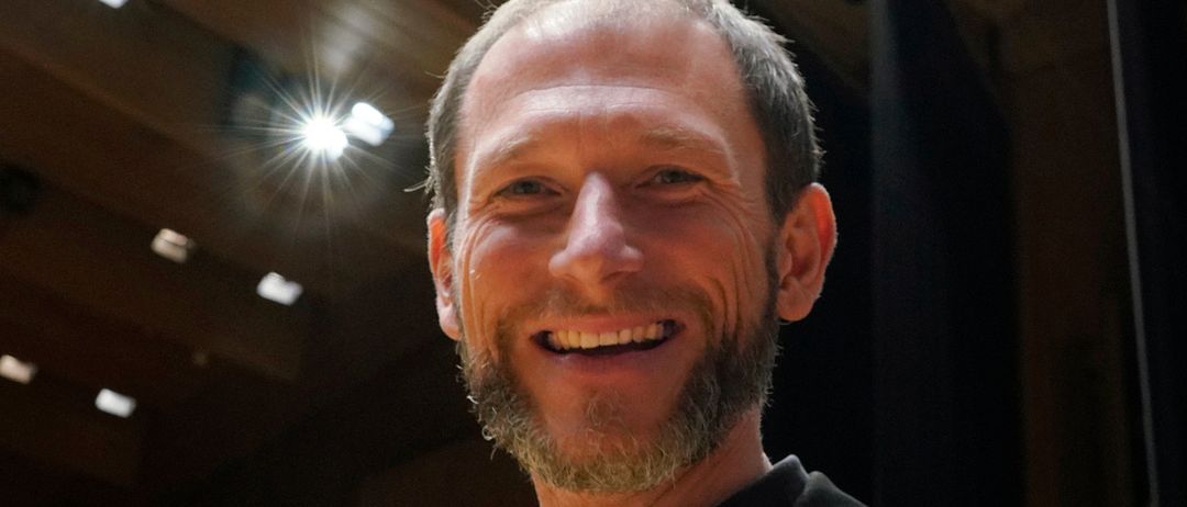 Ein Mann mit Bart und Lächeln steht vor einem hell beleuchteten Hintergrund. Er trägt ein schwarzes Hemd und einen Anzug.