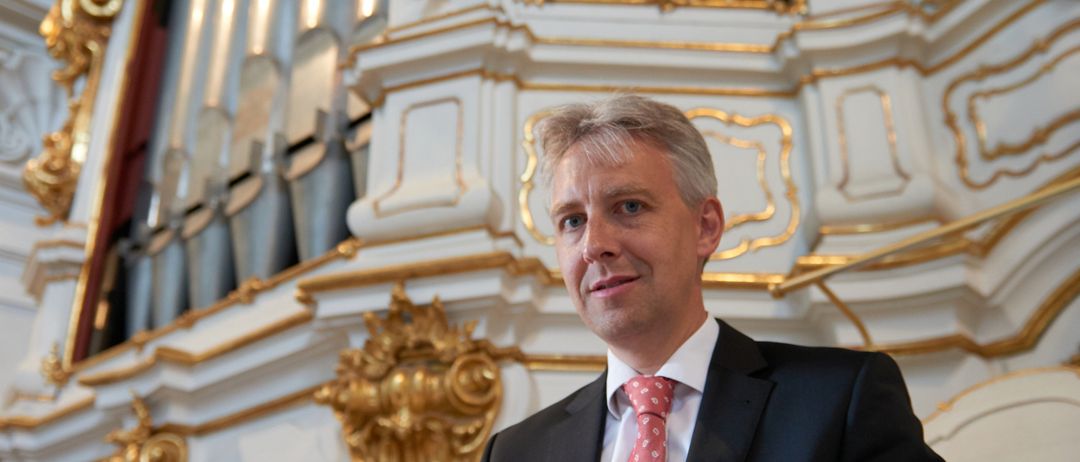 Ein Mann in einem schwarzen Anzug und roter Krawatte hält ein blaues Buch und liest vor einer kunstvollen Orgel.