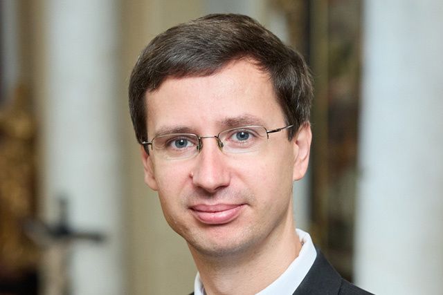 Ein Mann mit Brille und einem Anzugjackett lächelt in die Kamera. Der Hintergrund ist unschärfig.