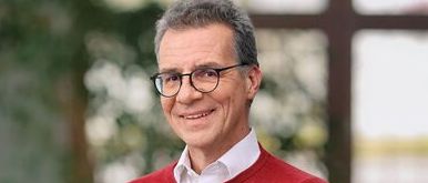 Ein lächelnder Mann mit Brille und rotem Pullover posiert für ein Foto vor einer unscharfen grünen Pflanze und einem Fenster.