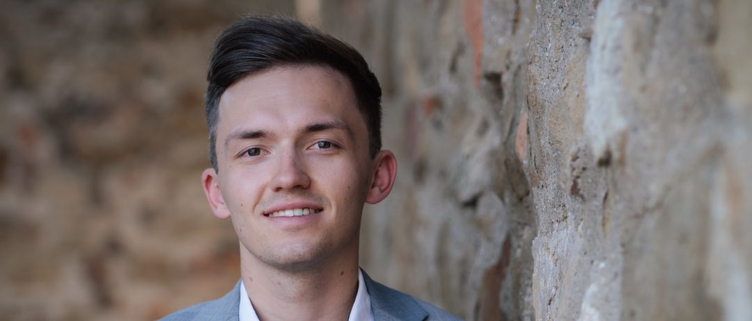 Ein junger Mann in einem grauen Anzug und weißem Hemd lächelt und lehnt sich gegen eine Steinmauer.