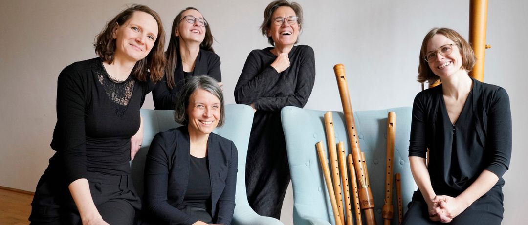 Fünf Frauen sitzen auf hellblauen Stühlen, lächeln und posieren für ein Foto. Dahinter steht eine Sammlung von hölzern Blockflöten auf einem weiteren Stuhl. Der Hintergrund ist weiß.
