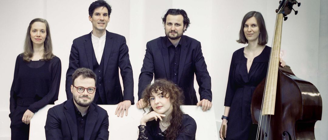 Eine Gruppe von Musikern in formeller Kleidung posiert für ein Foto. Zwei sitzen auf einer weißen Bank, während andere hinter ihnen stehen. Sie haben Instrumente wie ein Cello.