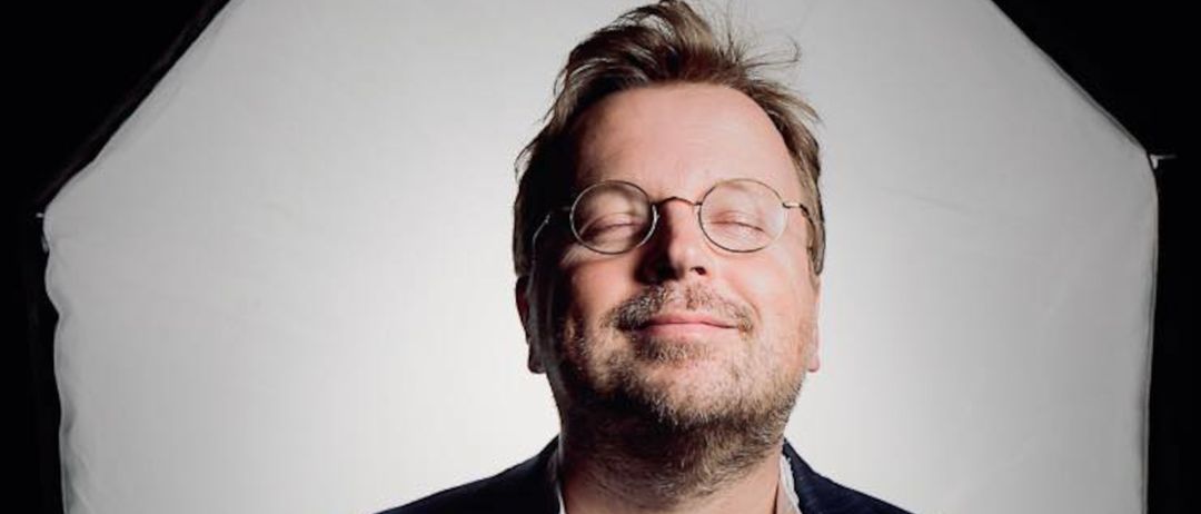 Ein Mann in einem schwarzen Anzug, weißem Hemd und Brille lächelt vor einem weißen Regenschirm.